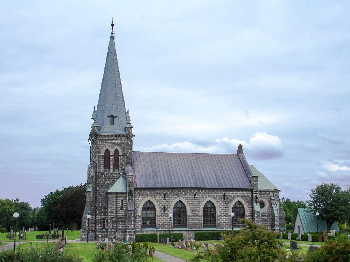 Trönninge kyrka i grå granit med hög spetsig tornspira, sedd från sidan över kyrkogården