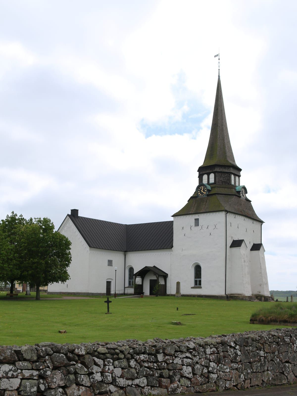 Veinge kyrka med vita murar och högt spånklätt torn med urtavlor, och en stenmur i förgrunden, i Veinge kyrkby